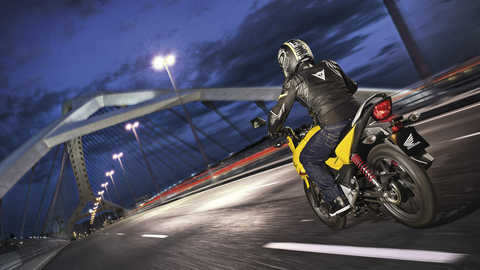 Vista traseira a tr&ecirc;s quartos de uma CB125F em amarelo Pearl Twinkle, virada para a direita, em estrada (cen&aacute;rio noturno em localiza&ccedil;&atilde;o urbana)