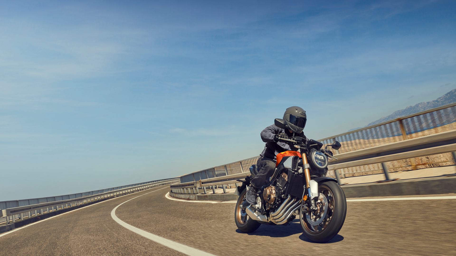 A Honda CB1000R motorcycle being ridden on a curved coastal road. The rider, wearing black gear, leans into the turn as the bike's distinctive round headlight and muscular frame are visible against a blue sky and ocean backdrop.