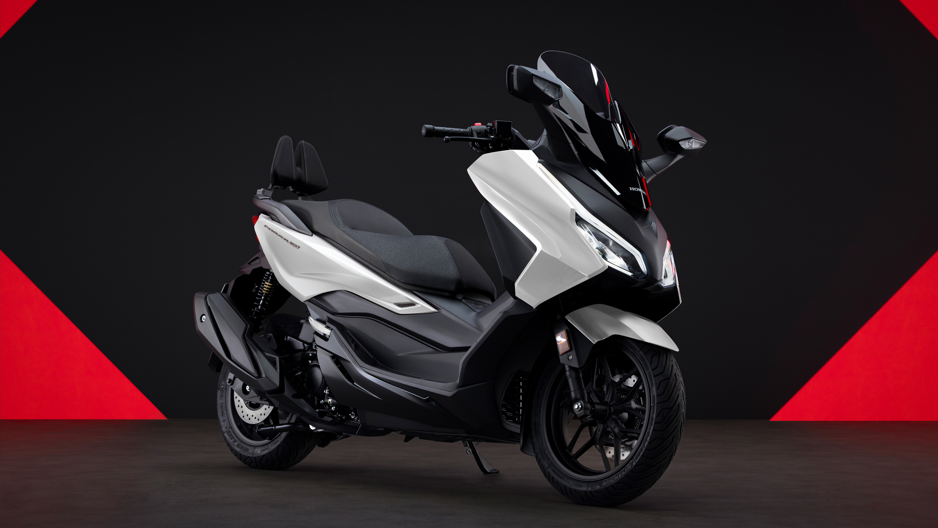 A white and black Honda Forza scooter positioned against a dark background with red triangular accents. The sleek maxi-scooter features a large windscreen, step-through design, and sporty styling.
