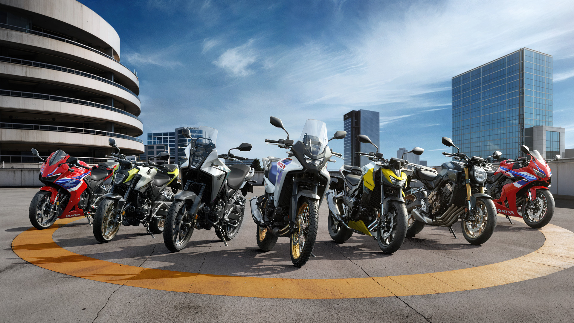 A lineup of various Honda motorcycle models, including sport bikes and adventure bikes, displayed on a rooftop parking area with modern city buildings in the background under a blue sky.