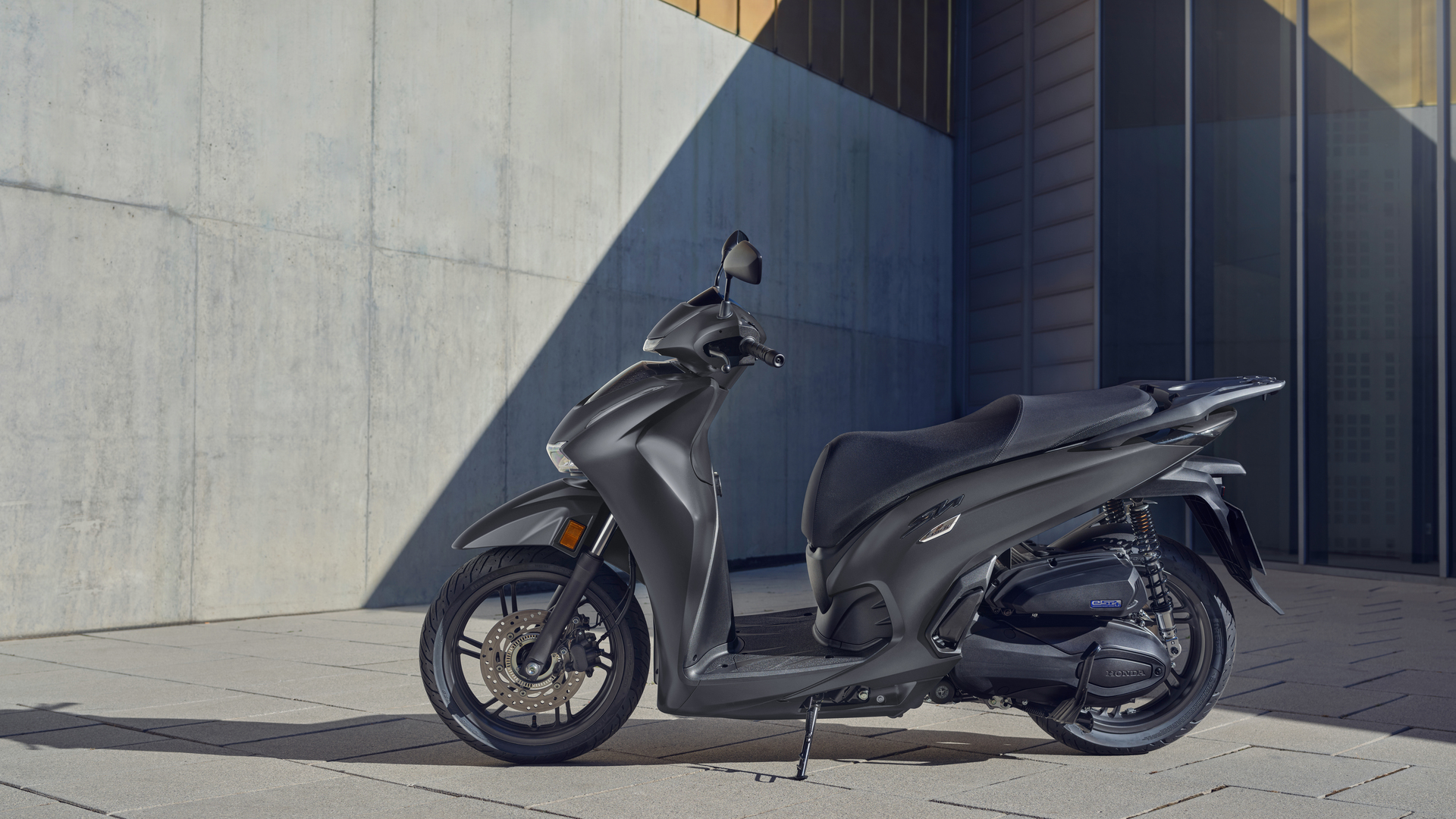 A sleek, modern Honda SH350i scooter in matte gray color parked on a sidewalk against a geometric concrete building backdrop. The scooter's streamlined design and large wheels are prominently displayed in profile view.