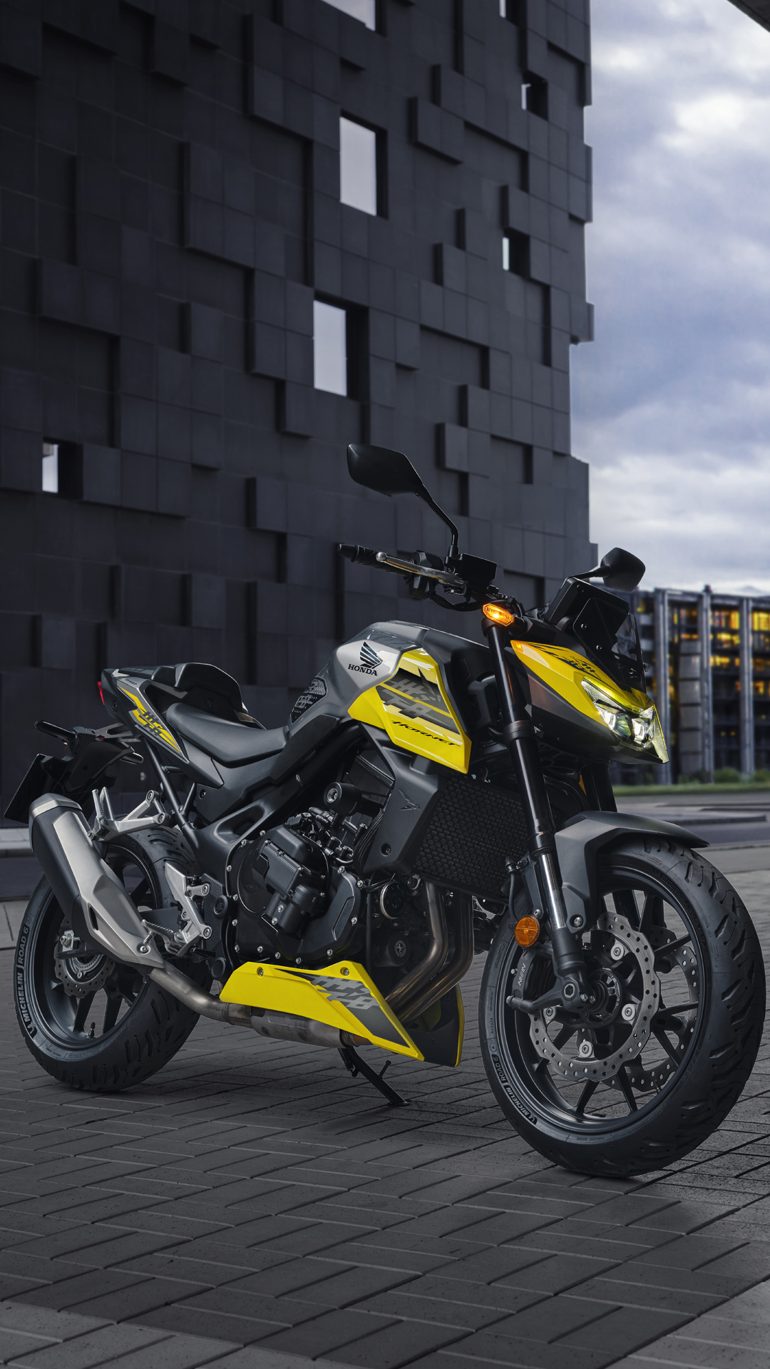 A yellow and black Honda CB500F motorcycle parked on a paved surface in front of a modern dark building with an unusual geometric facade. The sky is overcast, creating a moody urban atmosphere.
