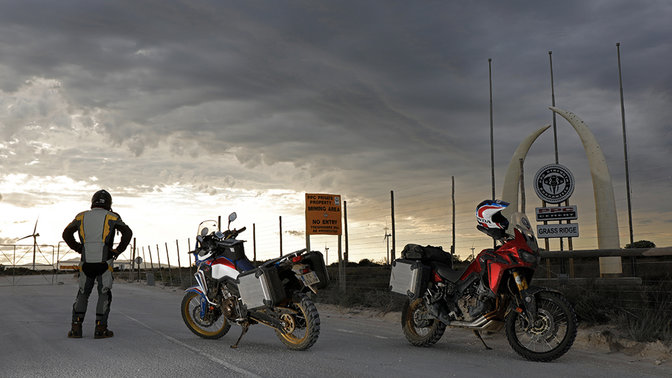 Piloto a olhar para o horizonte junto a duas Africa Twin estacionadas no alcatrão.