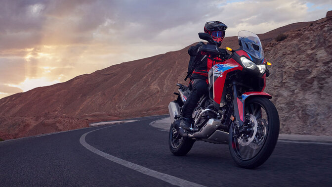 Modelo a conduzir a moto CRF1100L Africa Twin numa estrada num cenário de montanha.