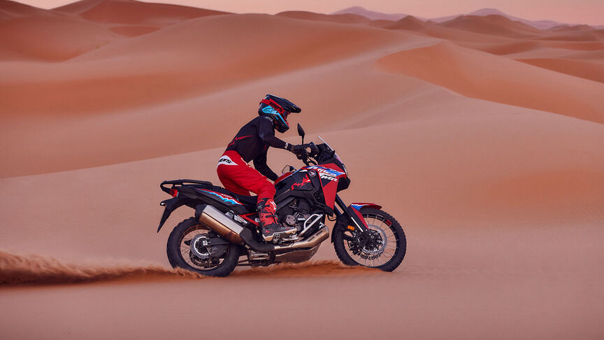 Modelo a conduzir uma mota CRF1100L Africa Twin num cenário de deserto.