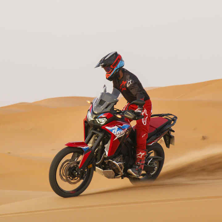 Modelo a conduzir a moto CRF1100L Africa Twin numa estrada num cenário de deserto.