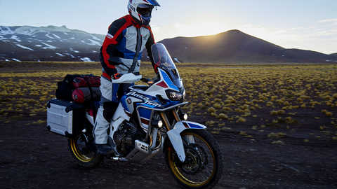 Vista dianteira de tr&ecirc;s quartos da Honda Africa Twin Adventure Sports com o condutor em p&eacute;, num cen&aacute;rio de montanha.