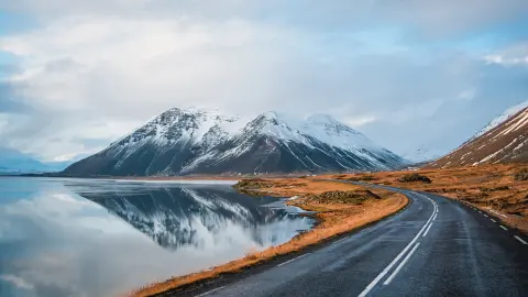 Fotografia panorâmica no inverno da estrada que percorre a costa do lago até às montanhas vulcânicas. Altos picos rochosos cobertos por uma camada de neve refletida sobre a superfície da água. Ponto de vista do condutor sobre a estrada de circunvalação, Islândia.
