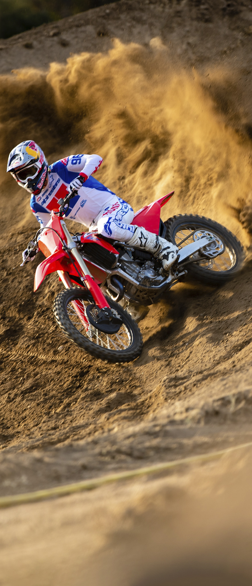 Um piloto de motocross, equipado com vestuário branco, azul e vermelho, inclina uma moto de motocross vermelha num talude de areia íngreme, levantando uma grande nuvem de terra ao virar na curva.