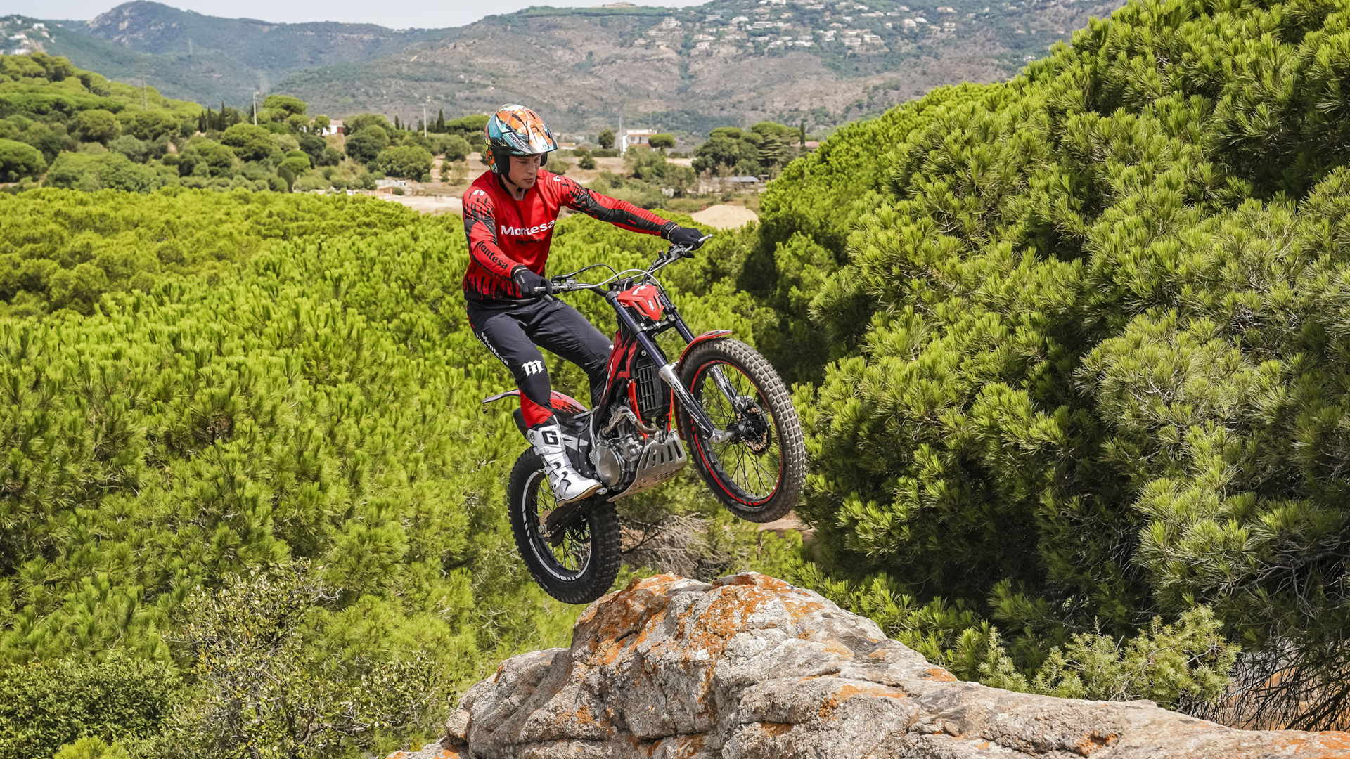 Motociclista a dar um salto com uma mota Montesa Cota 4RT 260R vermelha a partir de uma saliência rochosa, sobre uma paisagem pitoresca de um pinhal.