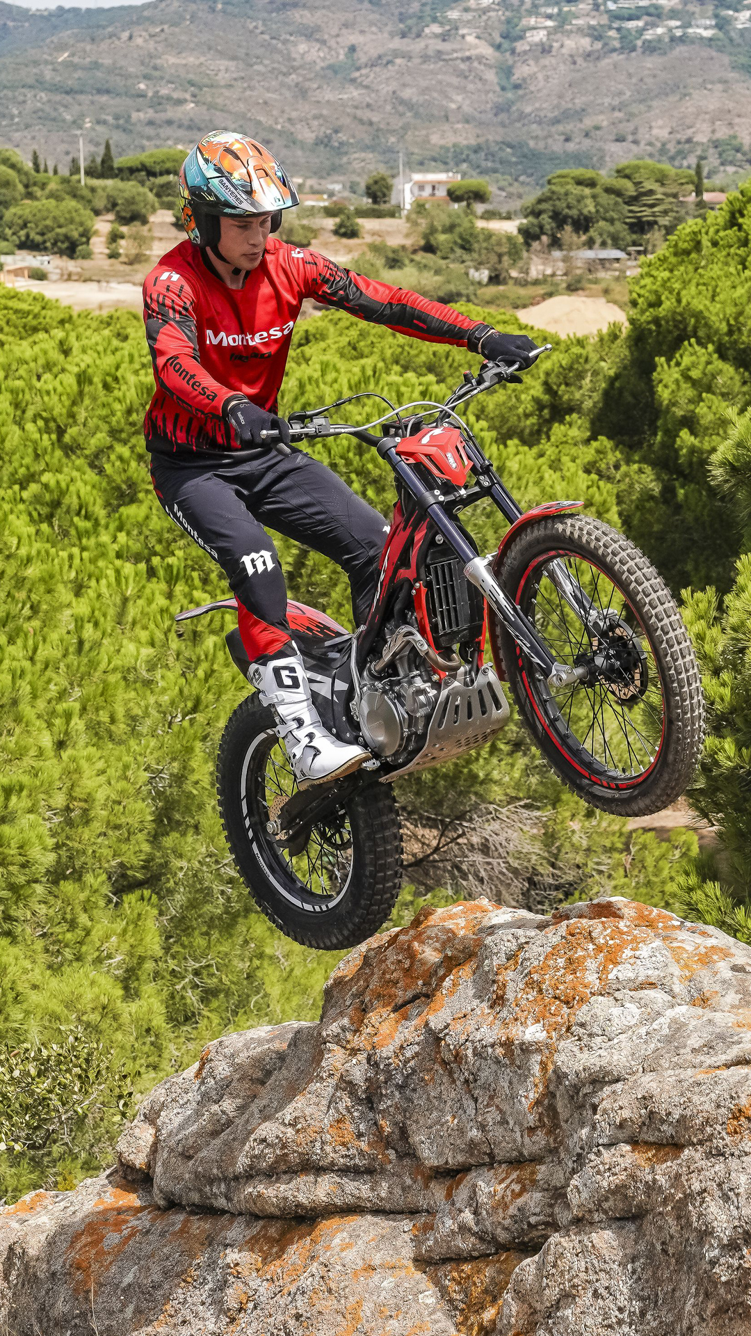 Motociclista a dar um salto com uma mota Montesa Cota 4RT 260R vermelha a partir de uma saliência rochosa, sobre uma paisagem pitoresca de um pinhal.
