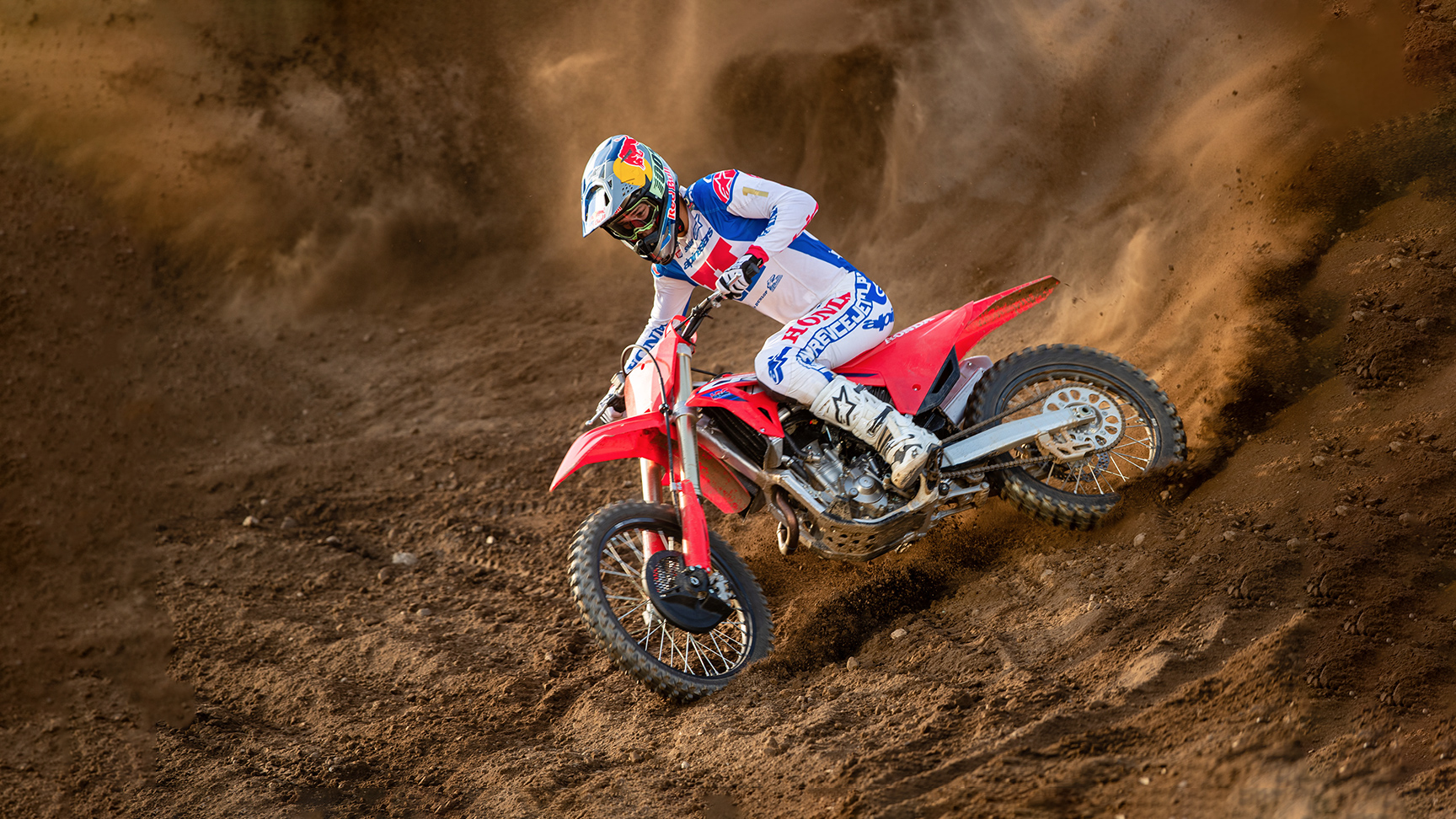 Um piloto de motocross, equipado com equipamento de corrida branca, azul e vermelha, faz uma curva acentuada numa moto de motocross vermelha, escavando uma pista arenosa e levantando uma impressionante nuvem de terra atrás de si.
