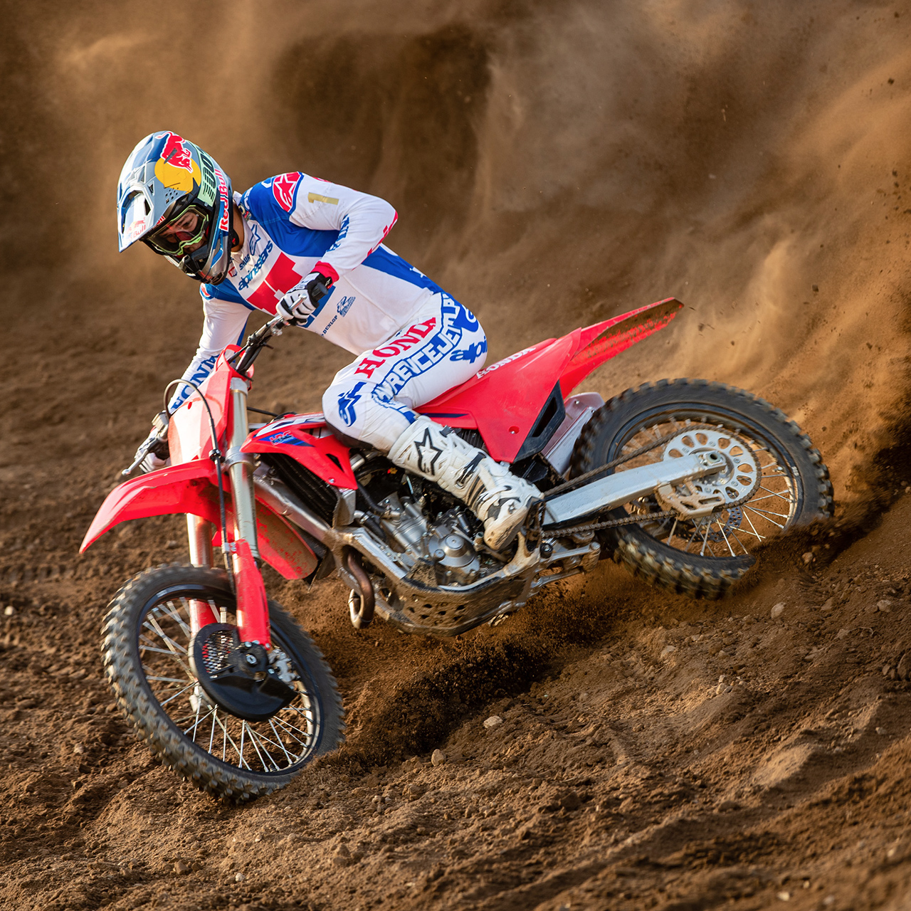 Um piloto de motocross, equipado com equipamento de corrida branca, azul e vermelha, faz uma curva acentuada numa moto de motocross vermelha, escavando uma pista arenosa e levantando uma impressionante nuvem de terra atrás de si.