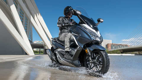 Vista dianteira a tr&ecirc;s quartos da Honda Forza 300, atravessando um curso de &aacute;gua.