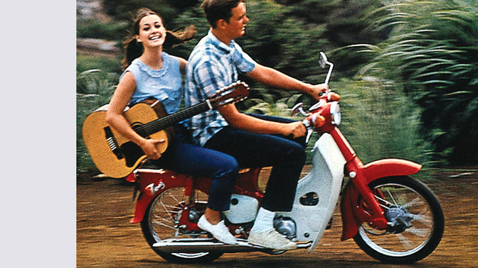 Mini-moto Honda com condutor e passageiro a transportar uma guitarra. Em estrada de terra batida. Virada para a direita.