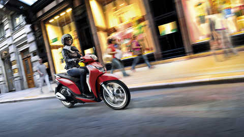 Perspetiva dianteira a três quartos da scooter Honda SH Mode cor-de-rosa com condutor. Virada para a direita (em estrada, noite).