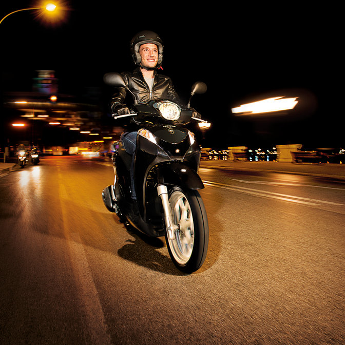 Perspetiva dianteira a três quartos da scooter Honda SH Mode preta com condutor. Virada para a direita (em estrada).