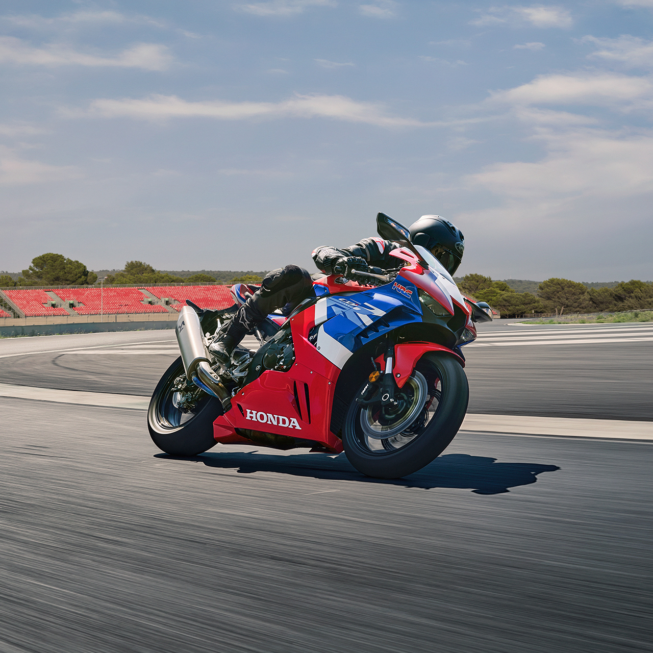 A rider in full black racing gear leans aggressively into a corner on a red, blue, and white Honda Fireblade sport motorcycle on a racetrack, with the asphalt blurred from speed and low greenery in the background.